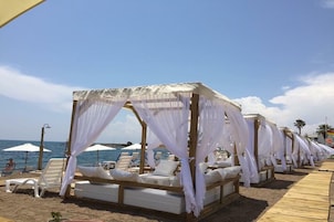 Un resort frente a la playa con hileras de camas y sillones de sombrilla blancos, un paseo de madera y sombrillas.