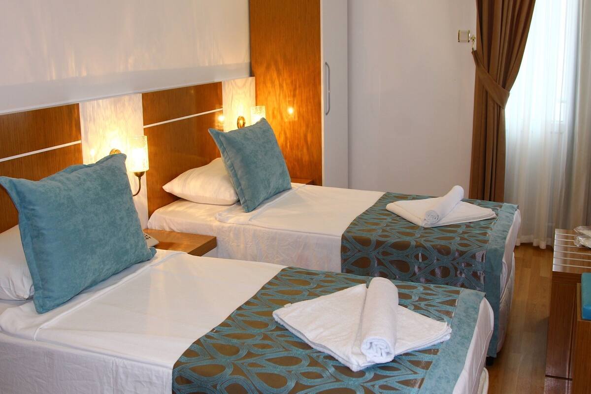 Een hotelkamer met twee bedden, elk voorzien van een beddekleed met dessin, kussens en een opgerolde handdoek. Tussen de bedden staat een nachtkastje met een lamp.