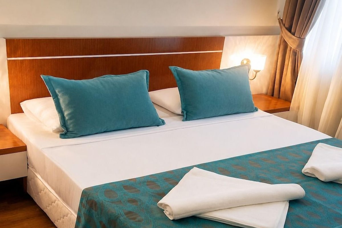 Une chambre d’hôtel avec un lit, deux oreillers, une table de chevet, une lampe et une fenêtre avec des rideaux.
