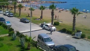 A coastal road with parked cars, a beach with umbrellas, and palm trees.