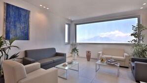A modern living room with a grey sofa, a white sofa, a coffee table, and a large window offering a view of the sky and mountains. - Relais Manfredi (Castellammare di Stabia)
