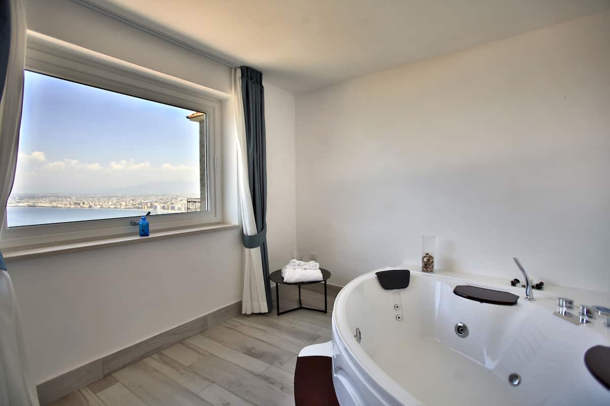 A modern bathroom with a large bathtub, a window with a view of a cityscape, and a small table with towels.
