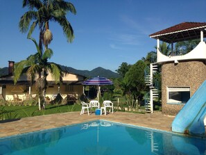 A swimming pool with a slide, surrounded by palm trees and a house with a patio. - Pousada Ecológica Cavalo de Pedra (Peruíbe)