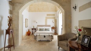 A bedroom with a stone archway, a bed with white bedding, a vase with flowers, and a framed picture on a side table.