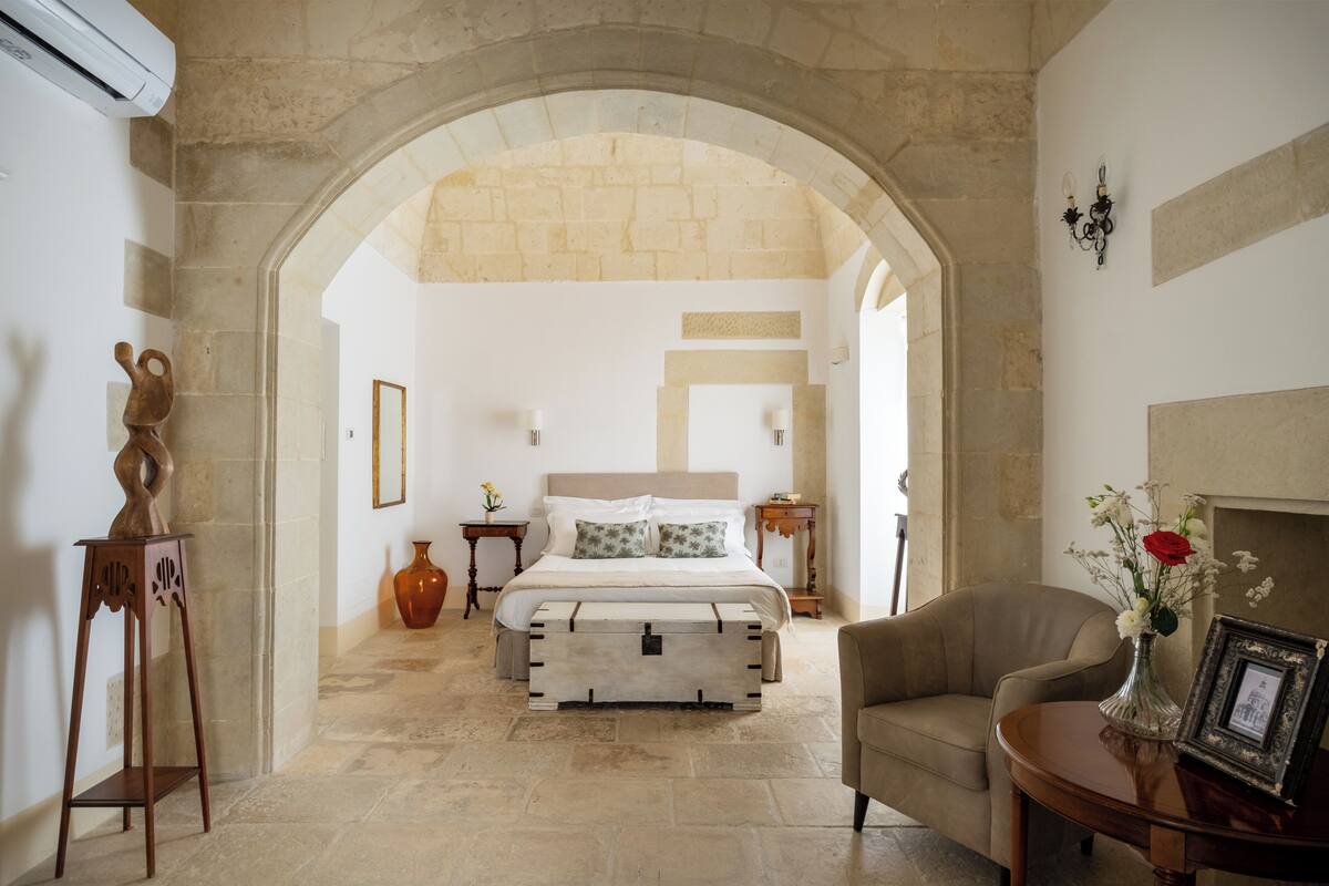 Une chambre avec une arche en pierre, un lit recouvert d’une literie blanche, un vase rempli de fleurs et un tableau encadré posé sur une table d’appoint.
