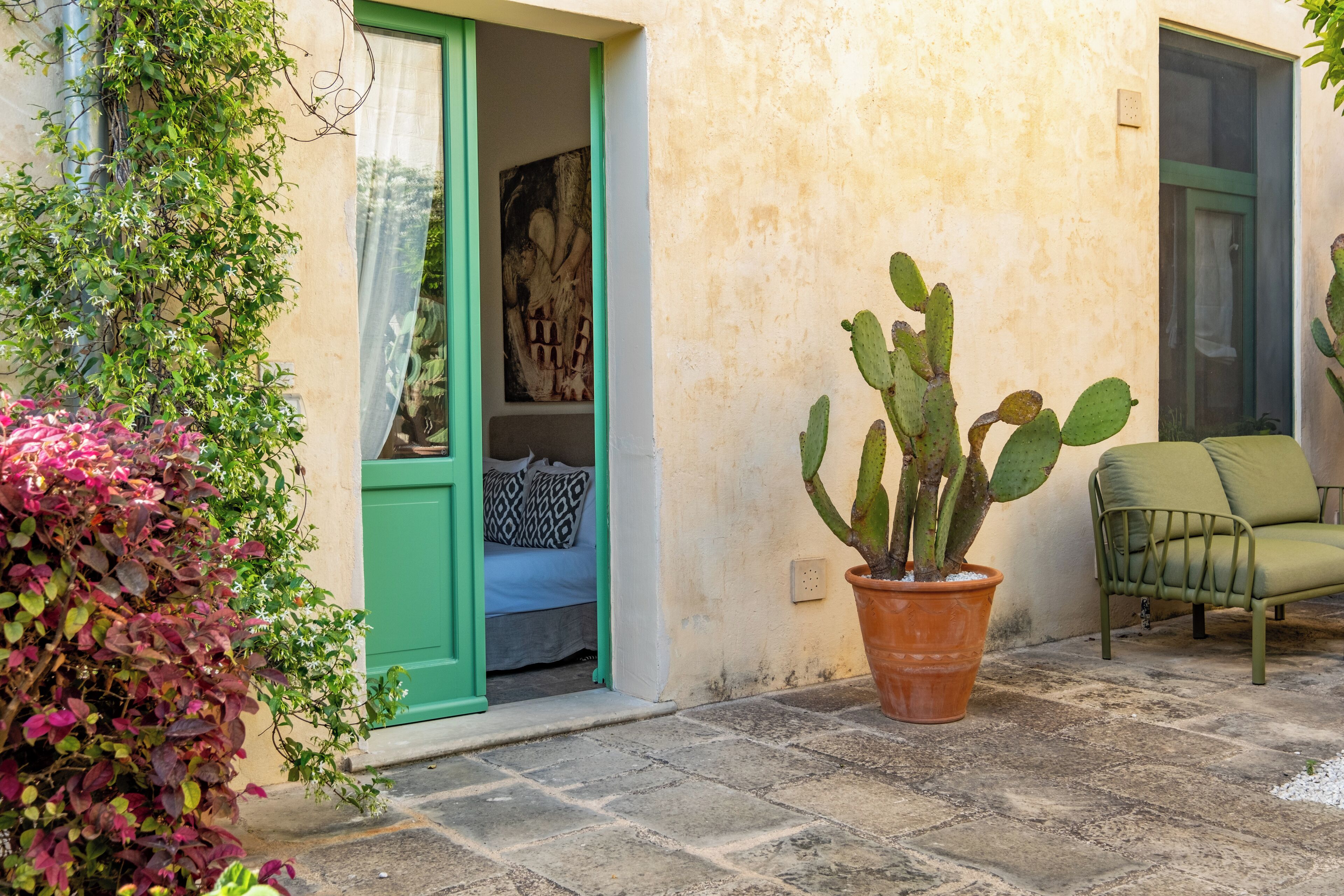 Un patio con una planta de cactus, una puerta verde, una cama con almohadas estampadas y una silla verde.