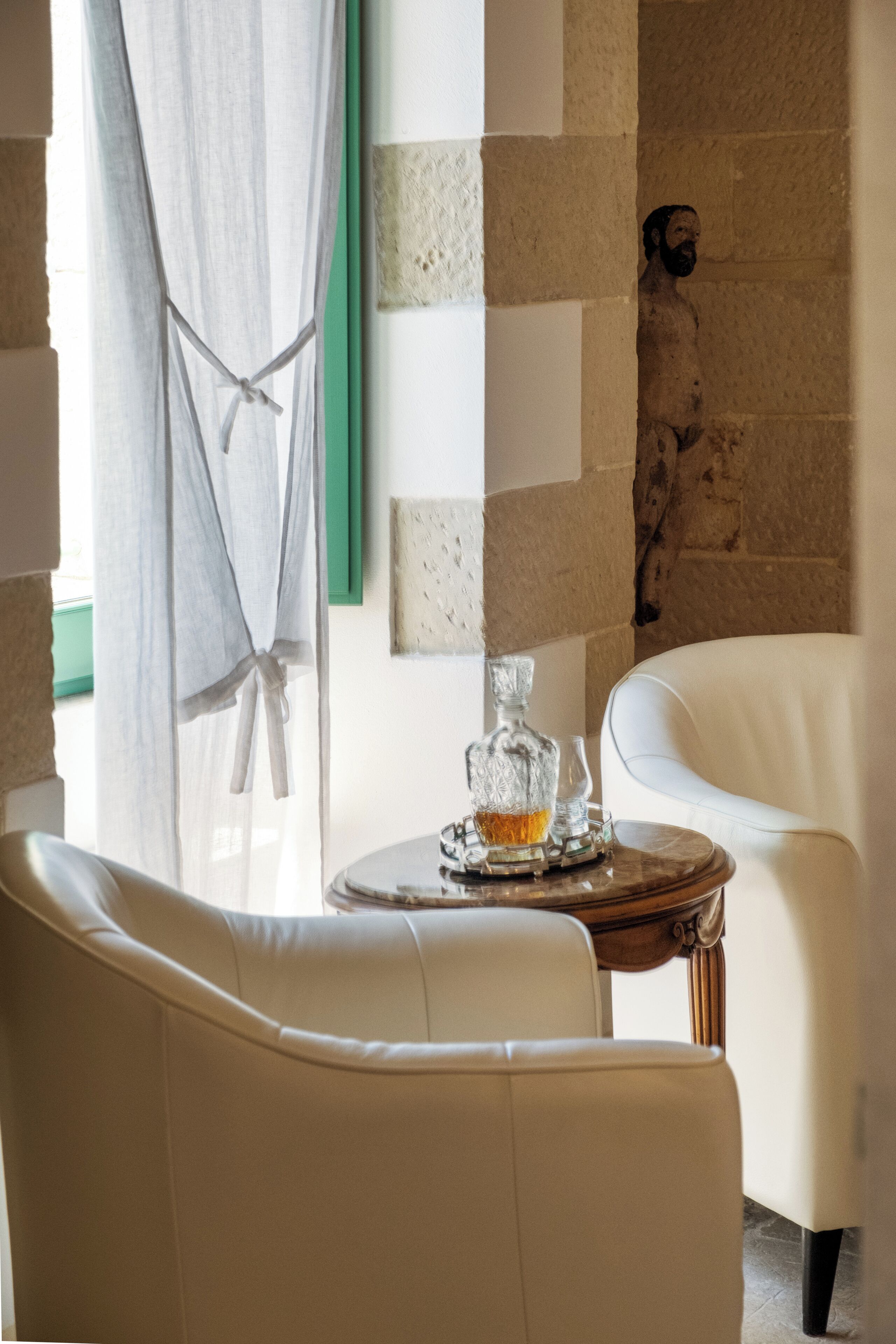 Ein Zimmer mit einem weißen Ledersofa, einem kleinen Holztisch mit einem Glaskarafell und einem Glas sowie einer Steinwand mit einer Statue.