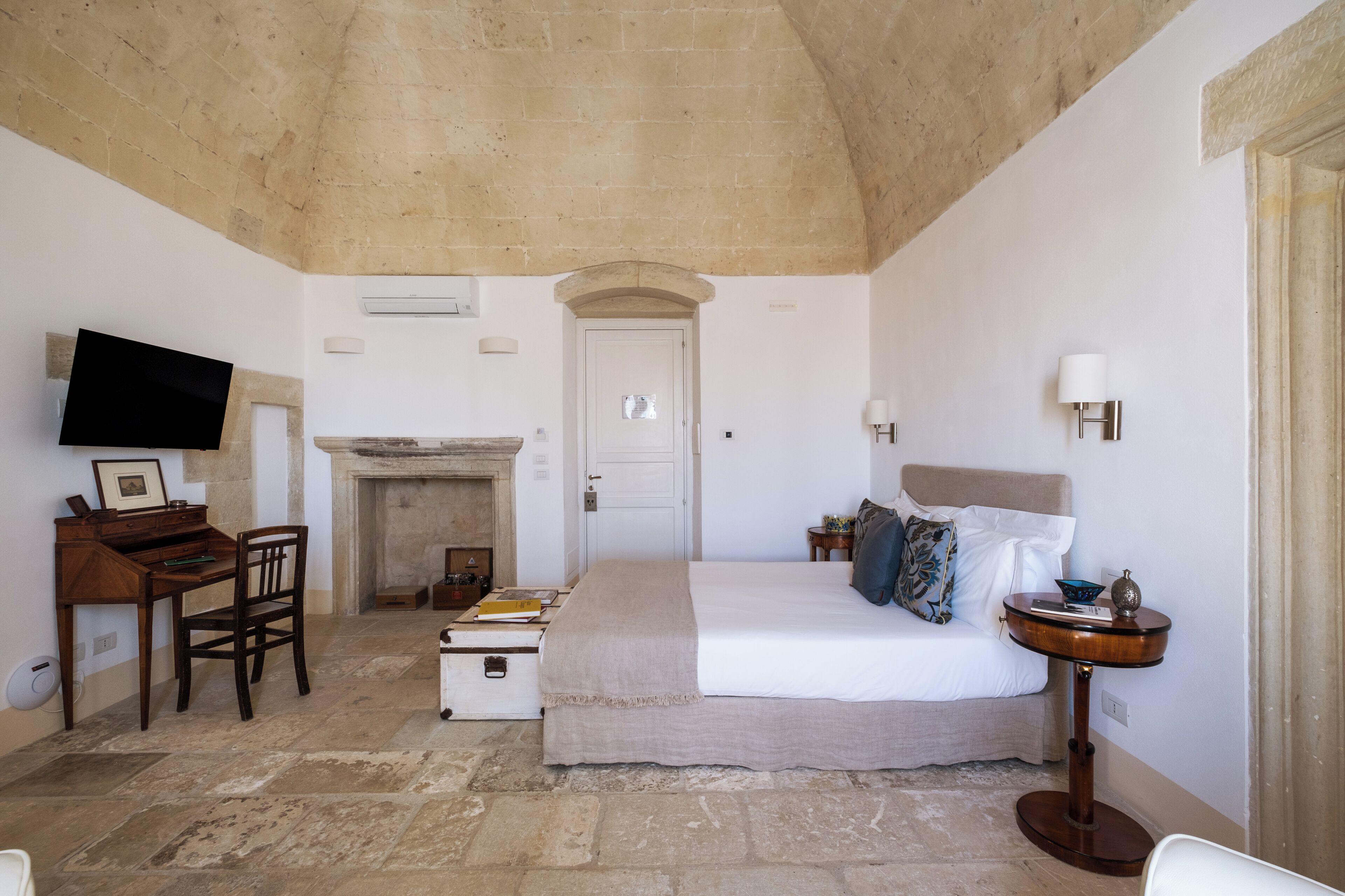 A bedroom with a bed, a desk, a television, a fireplace, and a small table with a lamp.