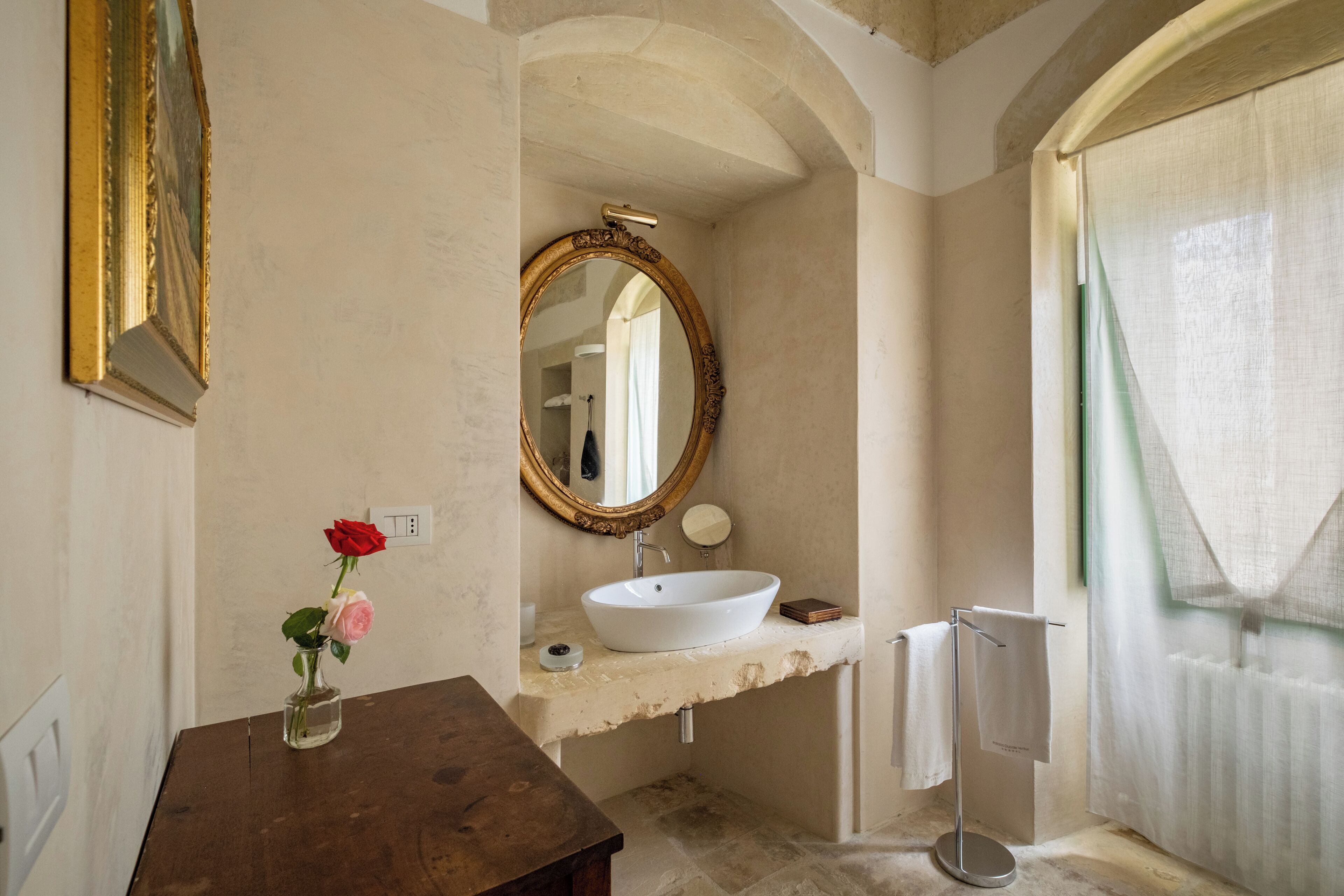 A bathroom with a white sink, a round mirror, a vase with flowers, and a towel rack.
