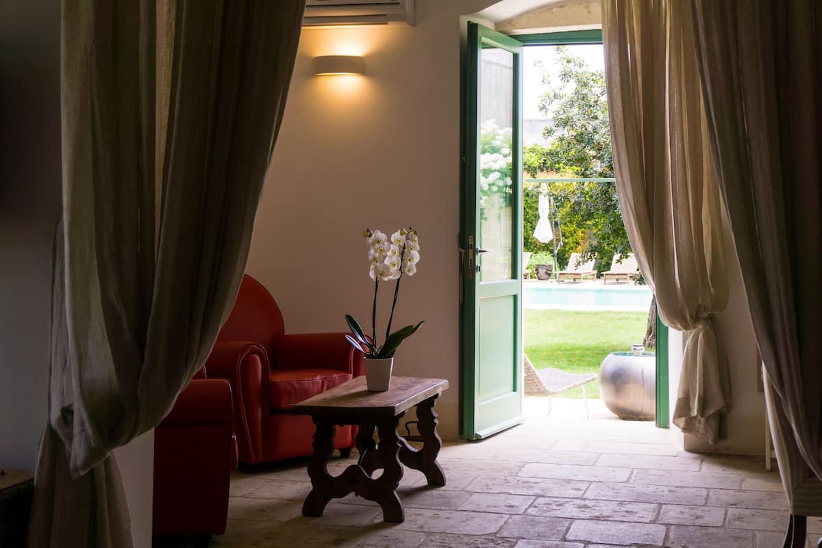 Une pièce avec un canapé rouge, une table en bois ornée d’une plante en pot, et une vue sur un jardin par une porte ouverte.