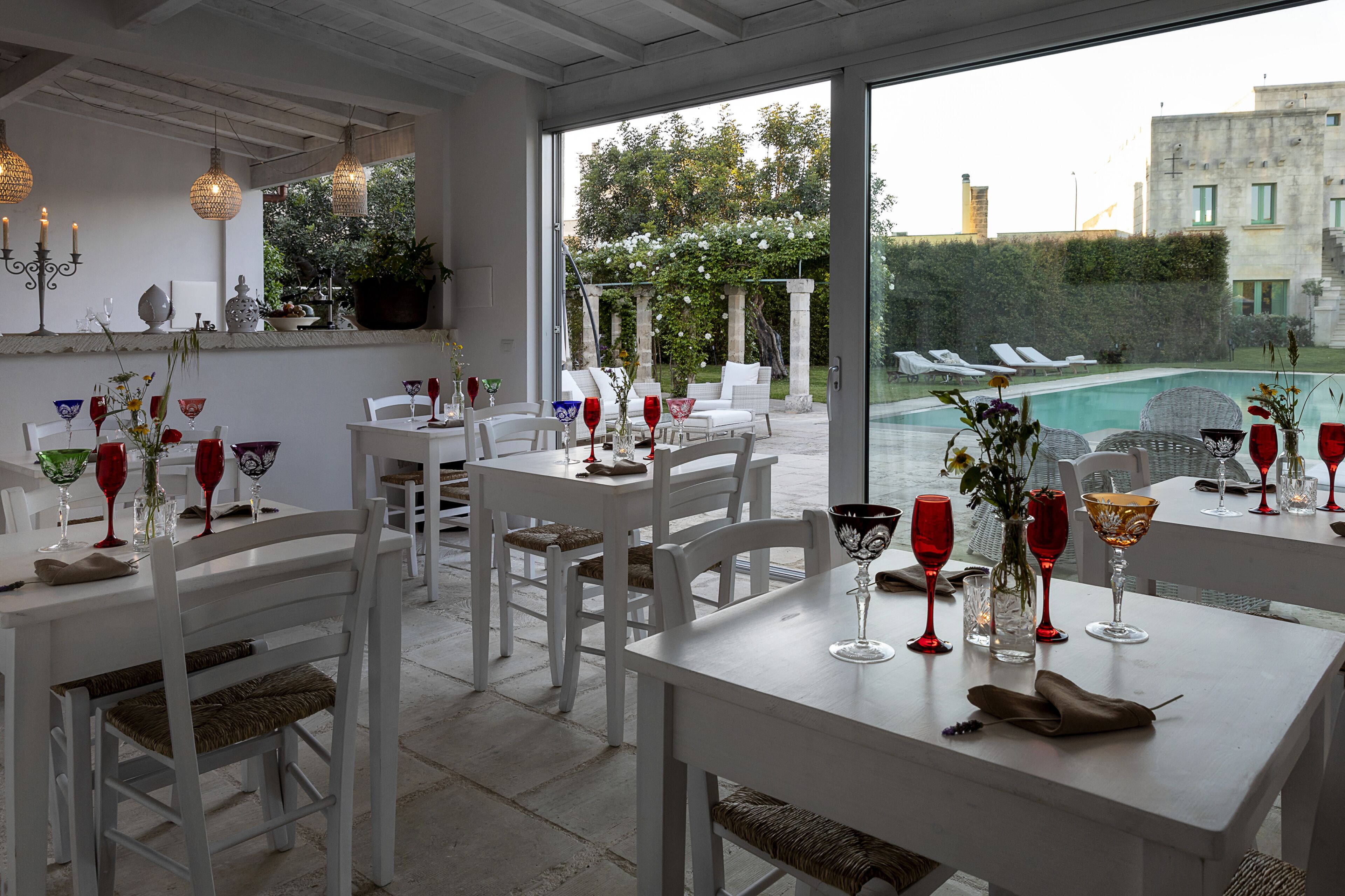 Un espace repas avec des tables et des chaises blanches, agrémenté de verres à vin rouge et de vases.