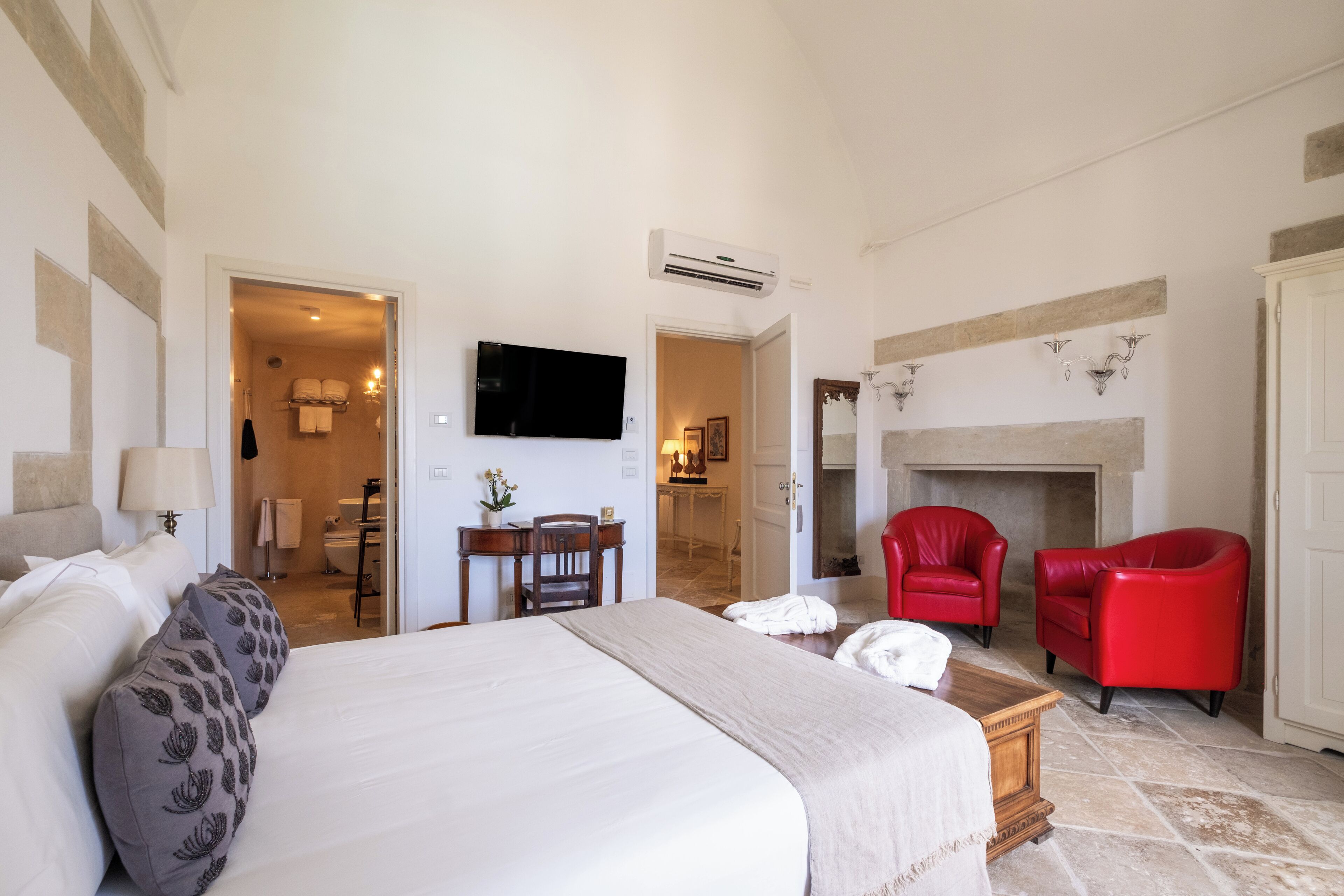 Une chambre d’hôtel avec un lit, un bureau, une télévision, des fauteuils rouges et une cheminée.