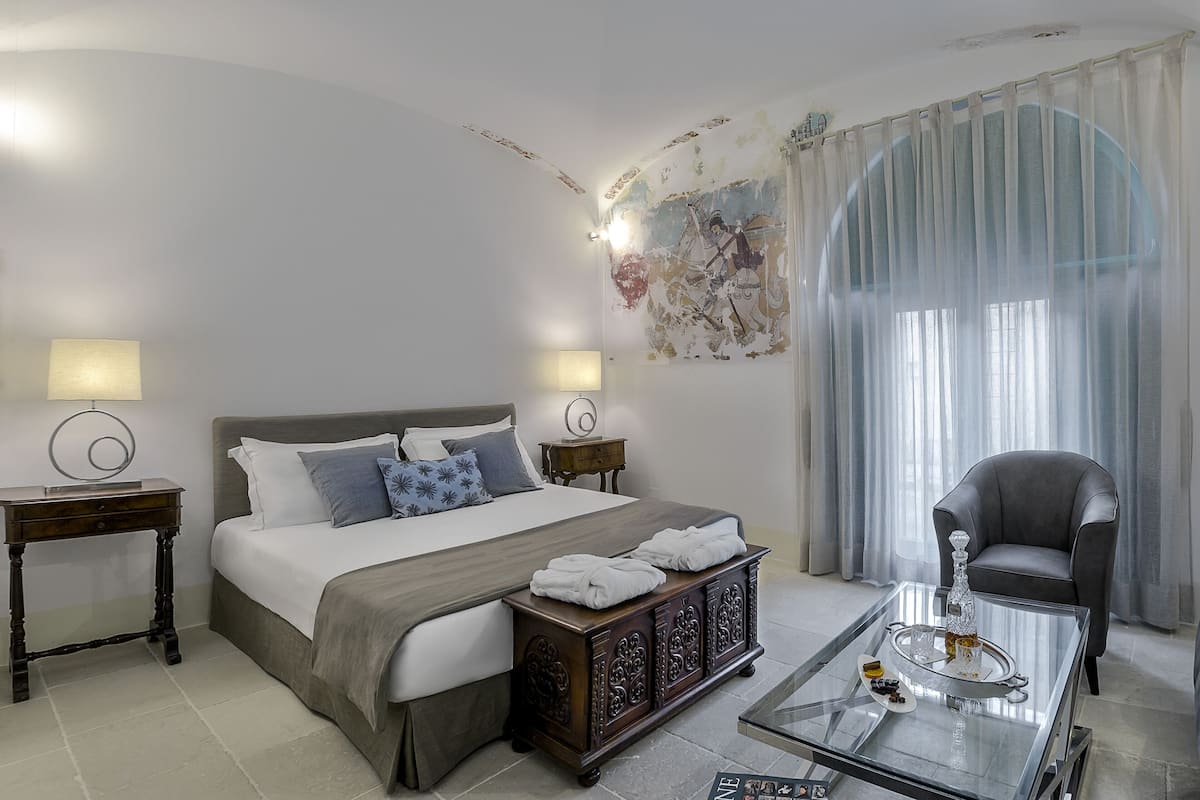 Une chambre d’hôtel moderne avec un grand lit, deux tables de chevet avec des lampes, une table basse en verre agrémentée d’un objet décoratif, un fauteuil et une grande fenêtre avec des rideaux légers.