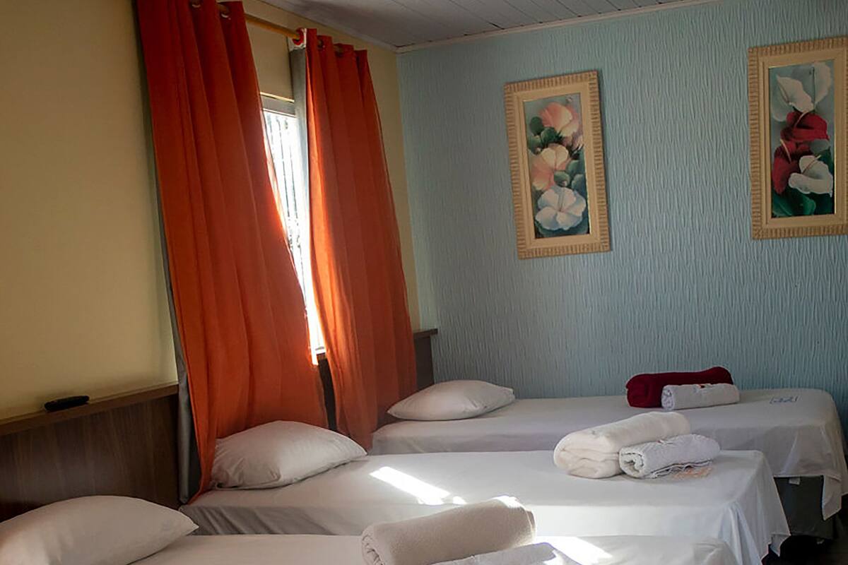 Quarto de hotel com quatro camas, cada uma com lençóis brancos e uma toalha. Há duas pinturas na parede, e cortinas laranja estão fechadas de um lado.