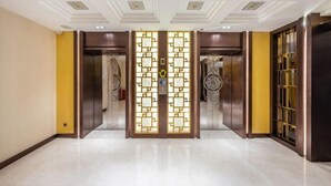 Un vestíbulo de hotel con una separación decorativa en el centro, dos ascensores y un mostrador de recepción.