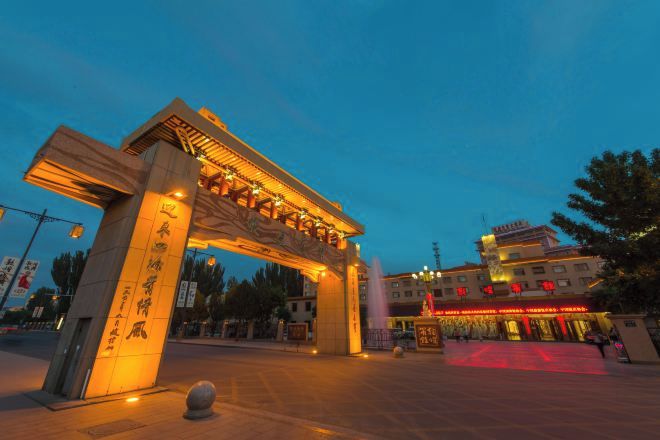 Un gran arco iluminado con caracteres chinos, una amplia plaza y un edificio con arquitectura tradicional al fondo.