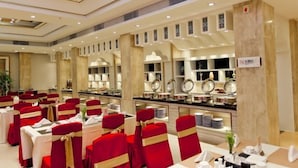 Un salón de banquetes con mesas dispuestas para un evento formal, con sillas rojas adornadas con cintas doradas, una zona de bufé con diversos platos y un diseño moderno en el techo.