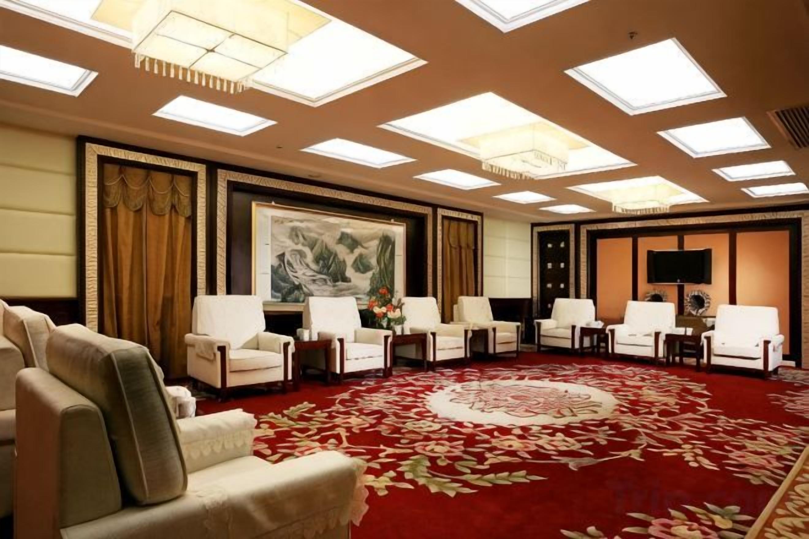 Une salle de conférence avec un tapis rouge, de nombreuses chaises, un grand tableau et des luminaires au plafond.