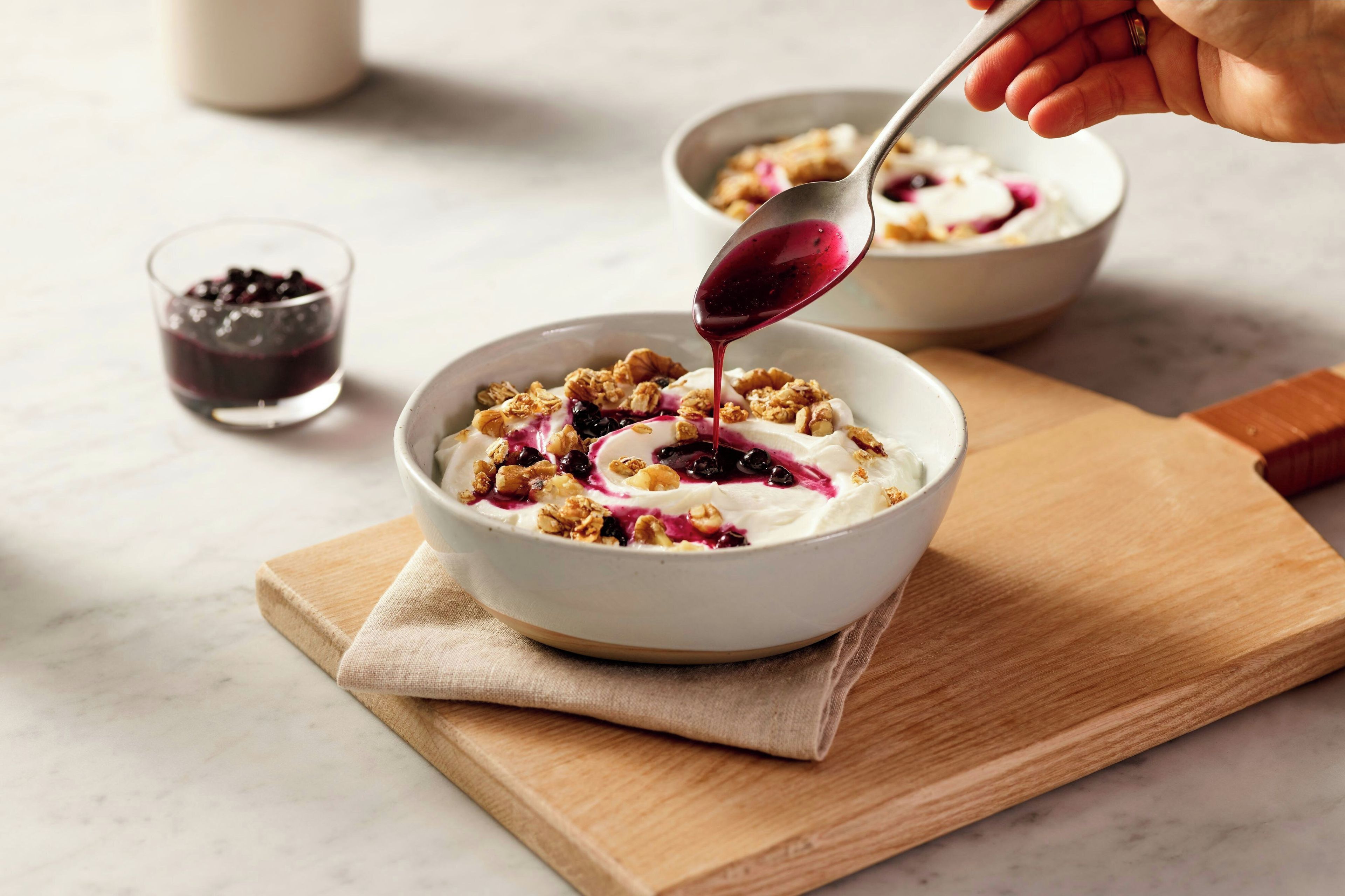 A bowl of yogurt topped with granola and berries, with a spoon pouring more berries into it.