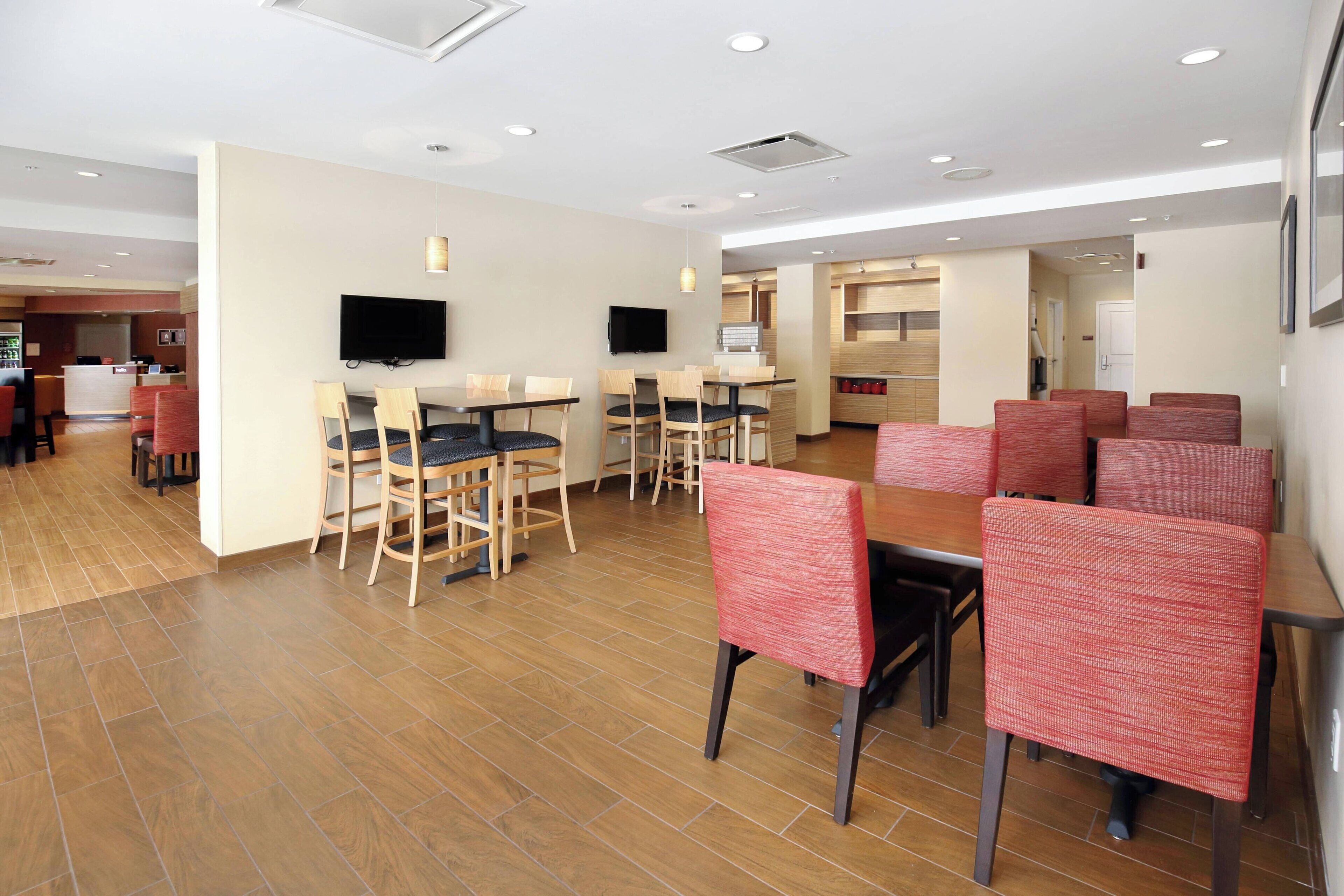 A hotel dining area with multiple tables and chairs, a television mounted on the wall, and a kitchen area in the background.