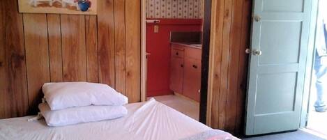 A wooden room with a bed, a painting of a beach scene, a kitchenette with red cabinets, and a door leading to another room.