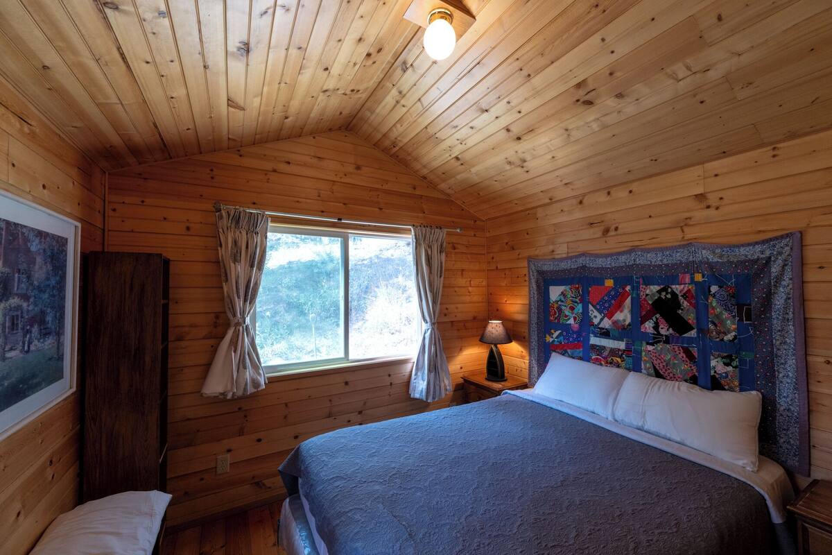 A wooden cabin bedroom with a bed, a window with curtains, a quilt on the wall, and a lamp on a nightstand.