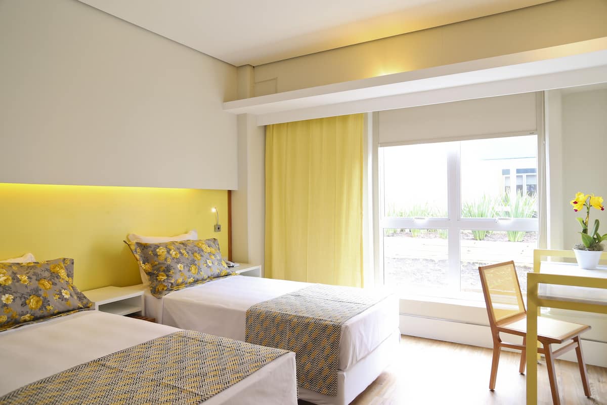 Habitación de hotel con dos camas, un escritorio y una silla. Se aprecia una ventana con cortinas amarillas y vistas al exterior.