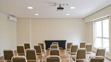 Uma sala de conferências com fileiras de cadeiras voltadas para uma mesa preta, um projetor e uma janela com persianas.