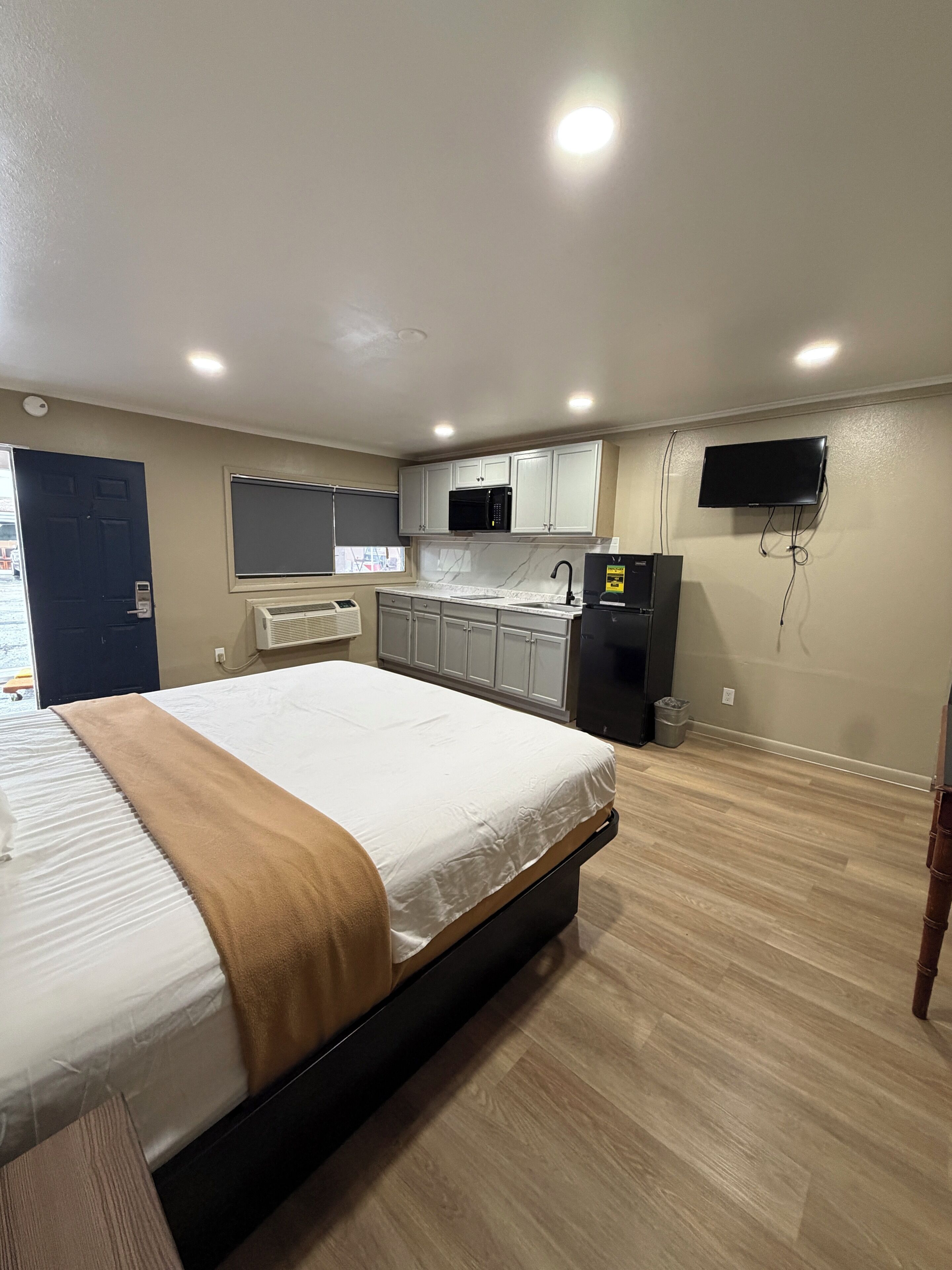 Quarto com cama, mesa de cabeceira, televisão, área de cozinha com armários e eletrodomésticos, e uma janela.