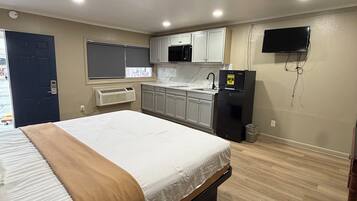 Quarto com cama, mesa de cabeceira, televisão, área de cozinha com armários e eletrodomésticos, e uma janela.