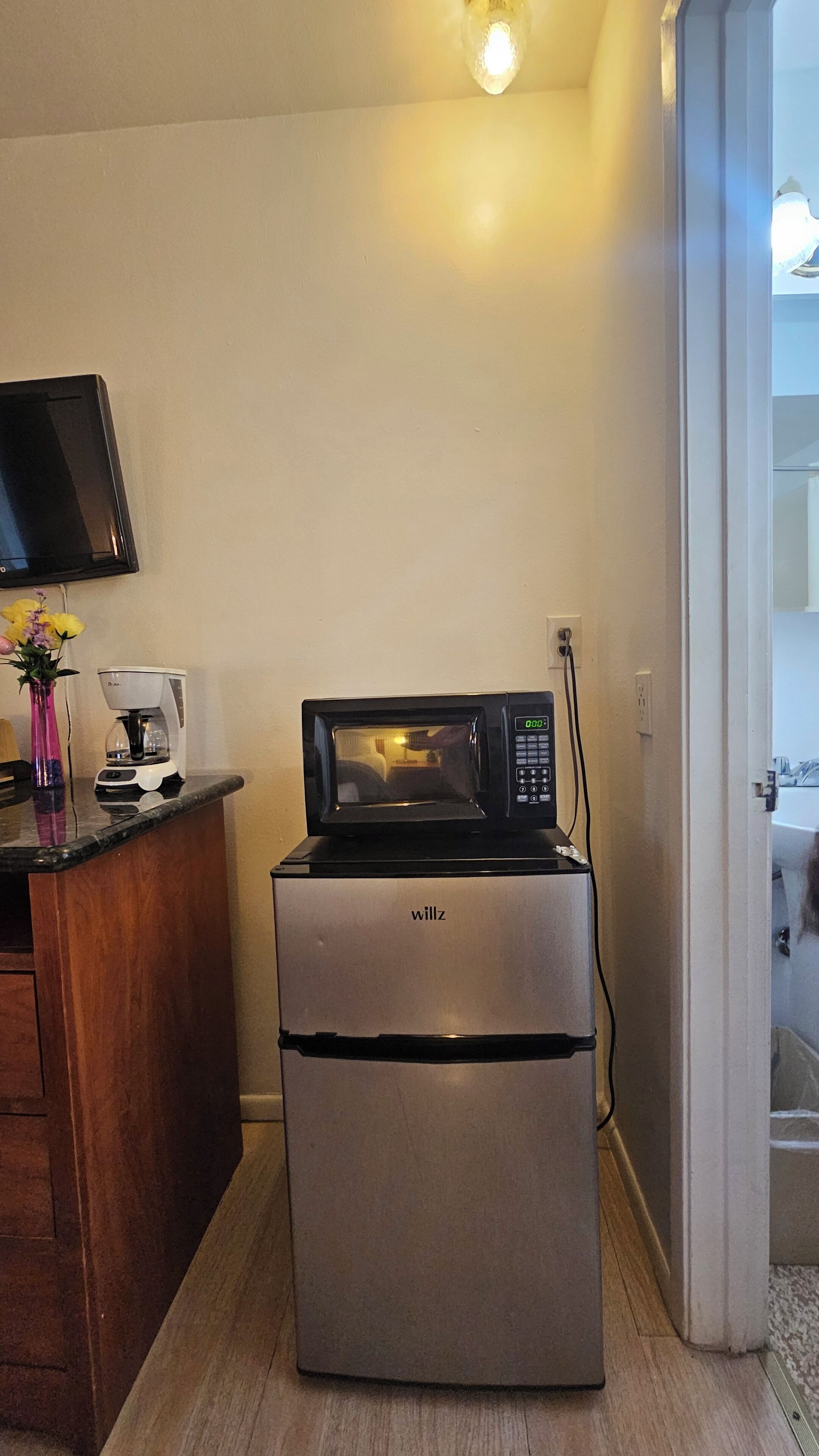 Et kompakt tekøkken med en mikrobølgeovn og en kaffemaskine på en bordplade, en lille vase med blomster, og et fjernsyn monteret på væggen.