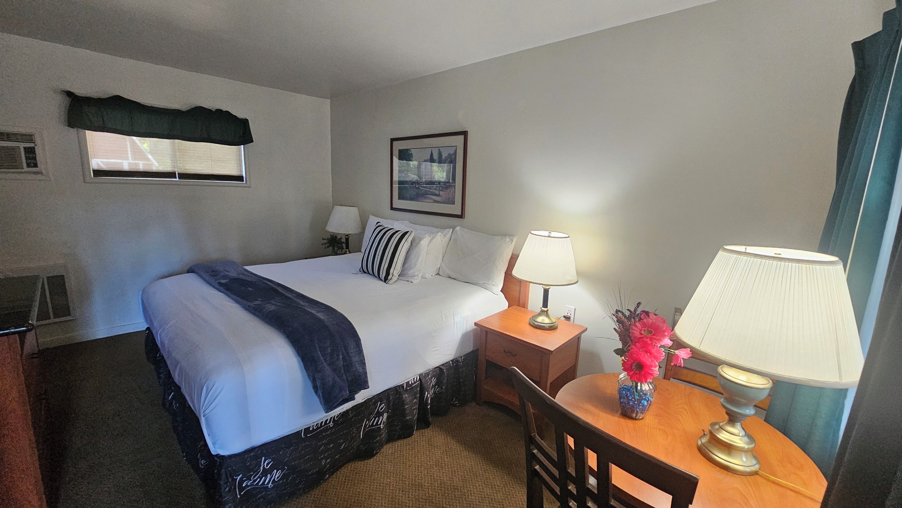 A hotel room with a bed, bedside tables, lamps, a chair, a window, and a painting on the wall.