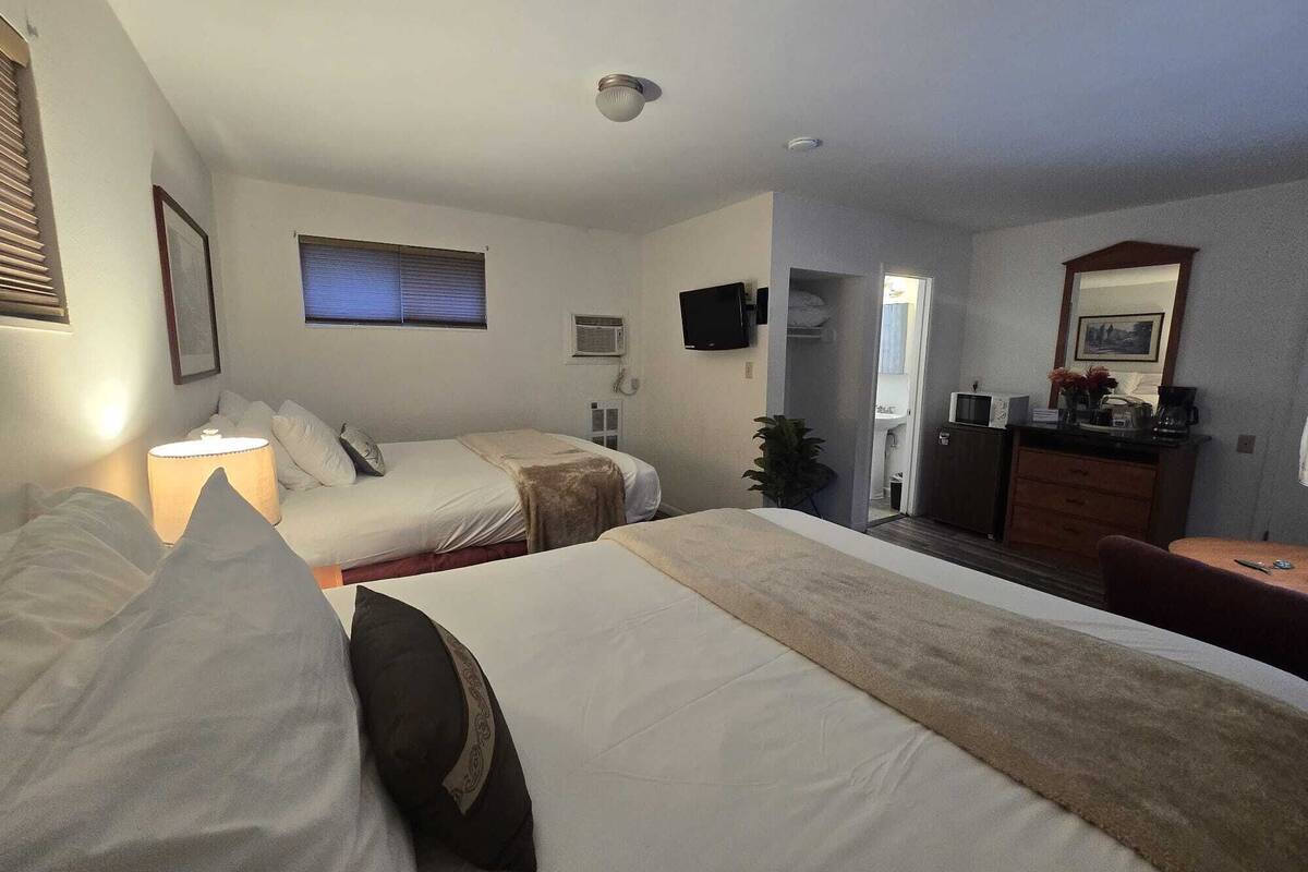 Quarto de hotel com duas camas, uma escrivaninha, cômoda, televisão e banheiro visível por uma porta aberta.