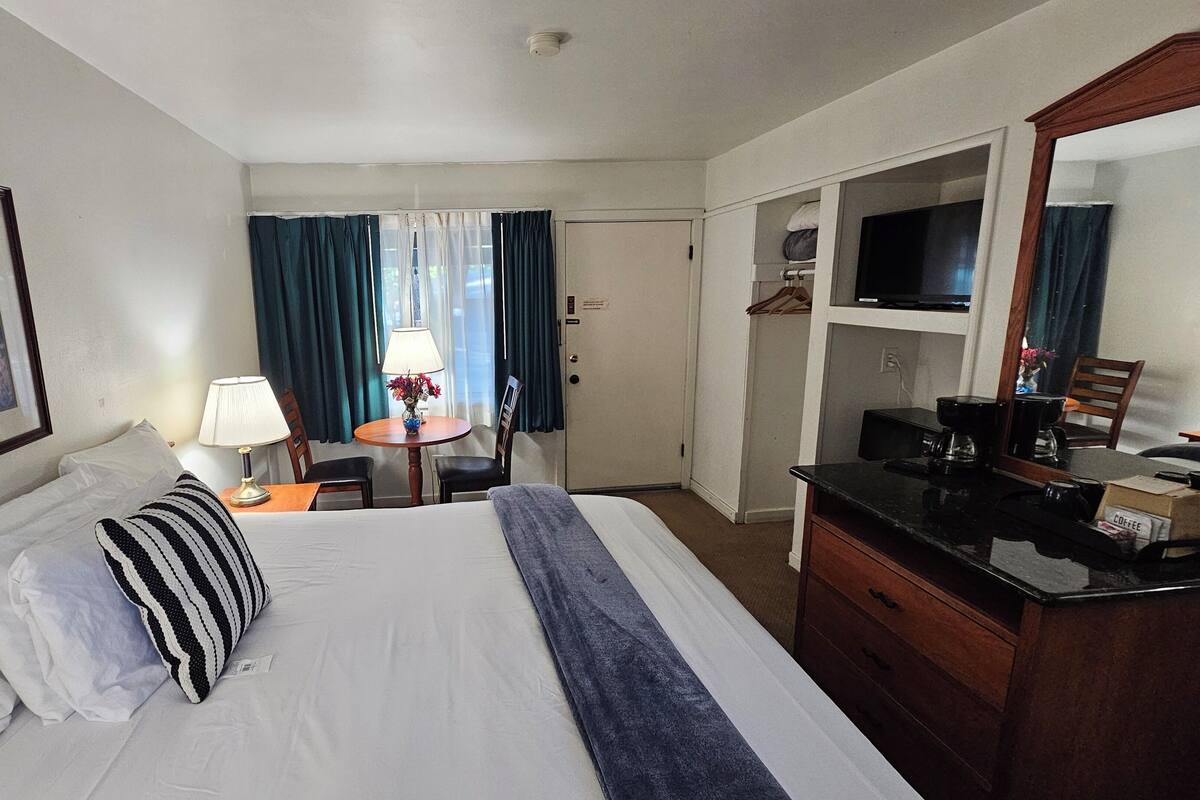 Quarto de hotel com cama, escrivaninha, cadeira, televisão e guarda-roupa.