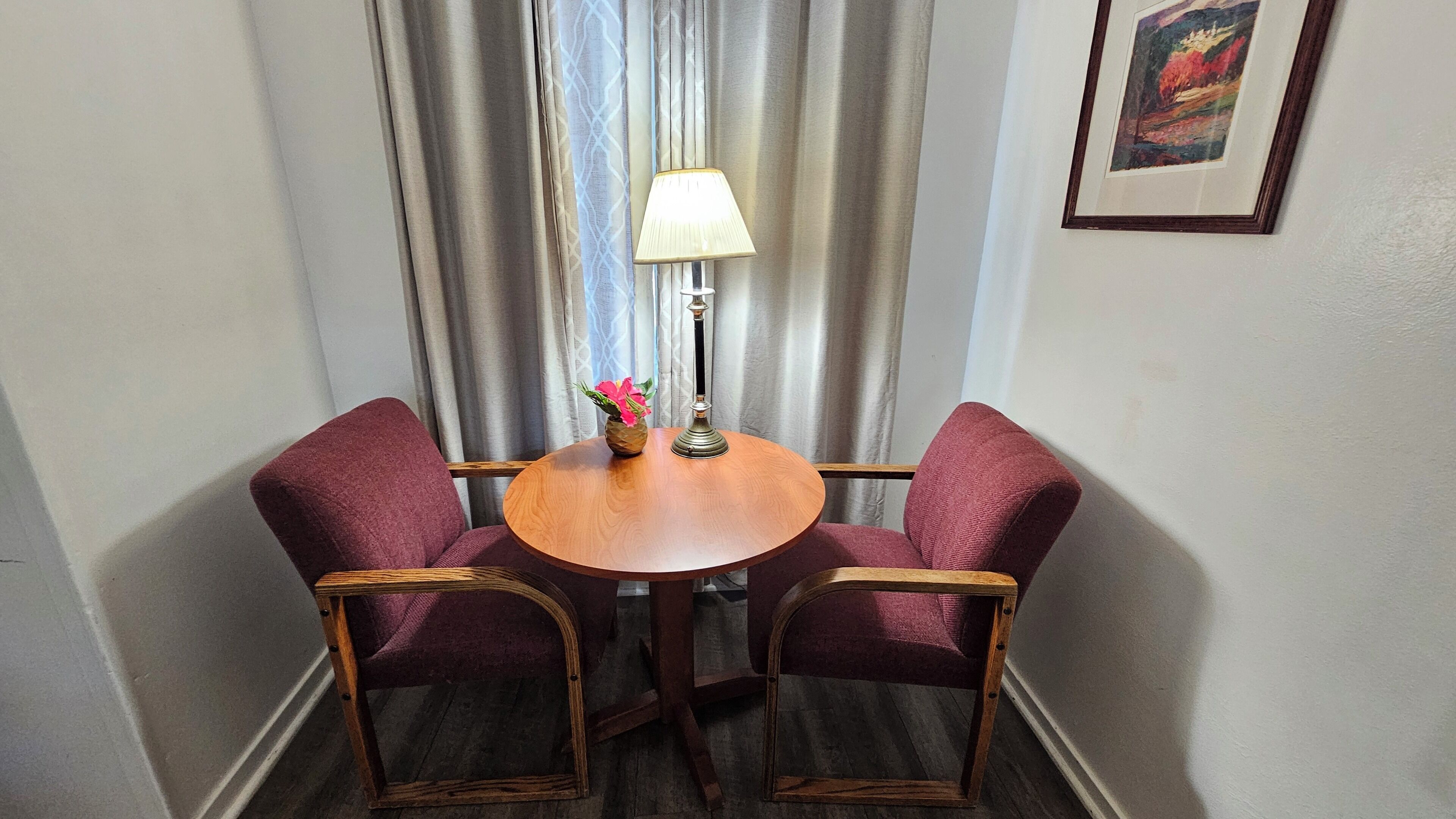 Um quarto pequeno com uma mesa redonda de madeira, duas cadeiras estofadas, um abajur e um quadro na parede.
