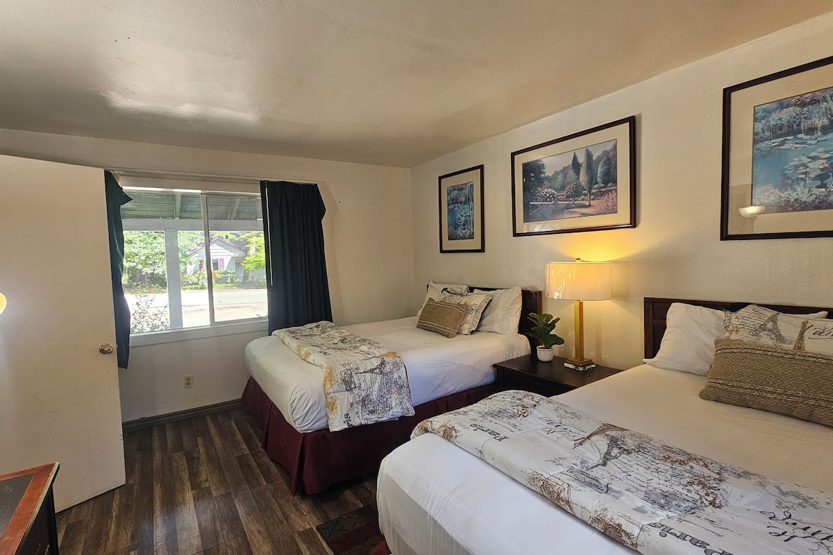 Quarto de hotel com duas camas, janela com cortinas, escrivaninha com luminária e quadros na parede.