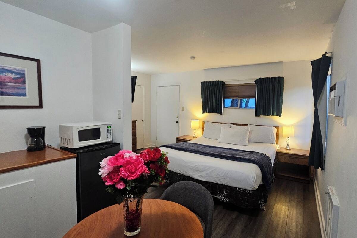 Quarto de hotel com cama, luminárias laterais, micro-ondas, cafeteira e um vaso com flores.