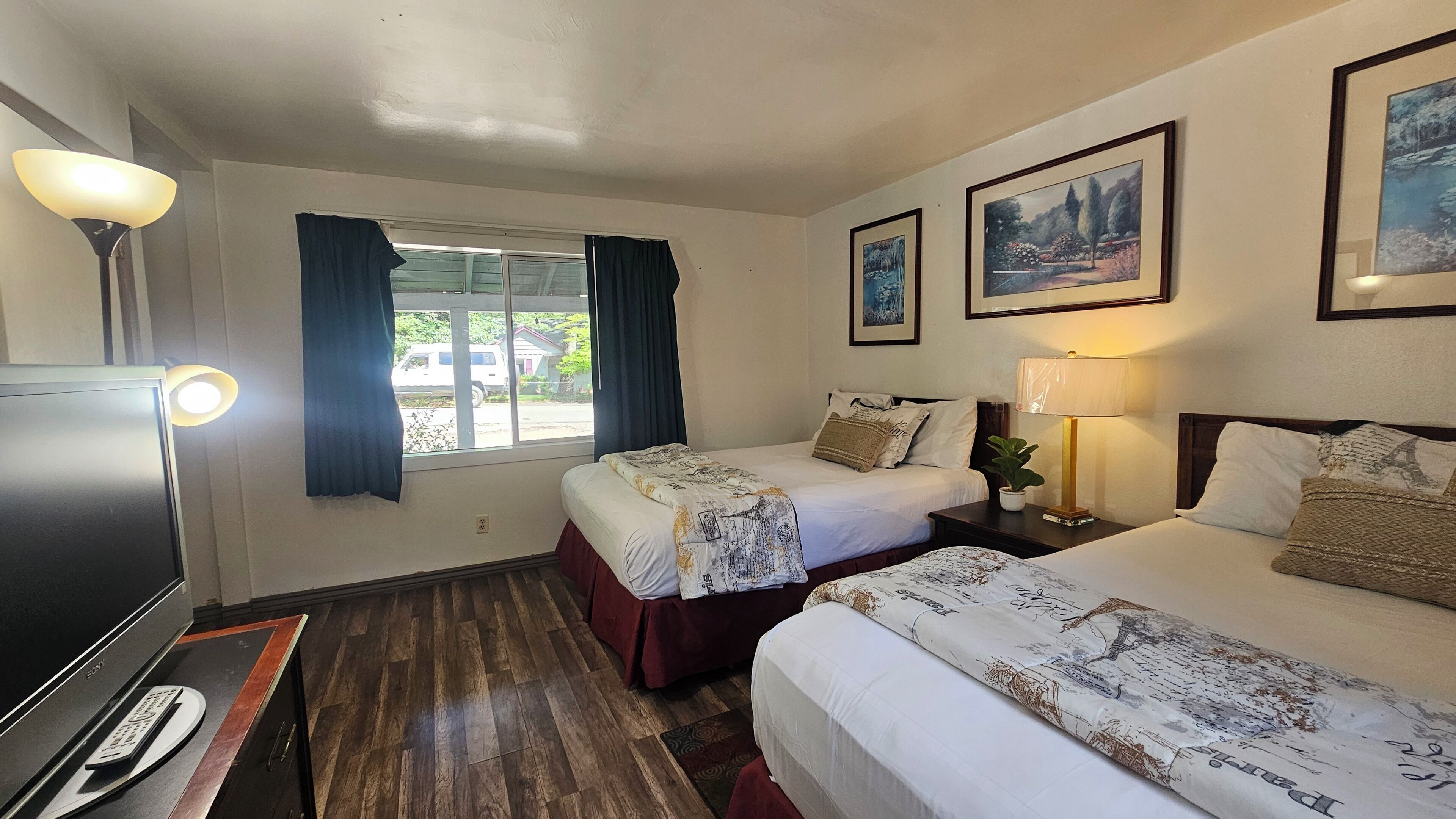 A hotel room with two beds, a TV, a window with curtains, and framed pictures on the wall.