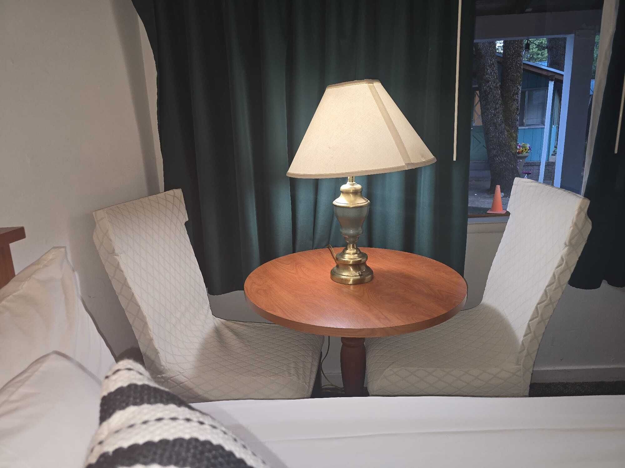 Une chambre d’hôtel comprenant un lit, deux fauteuils rembourrés, une petite table en bois avec une lampe et une fenêtre avec des rideaux.