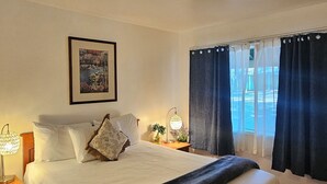 A neatly made bed with a white comforter and a dark throw blanket, a framed picture on the wall, a bedside lamp, and a window with dark curtains. - Cedar Lodge Motel (Dunsmuir)