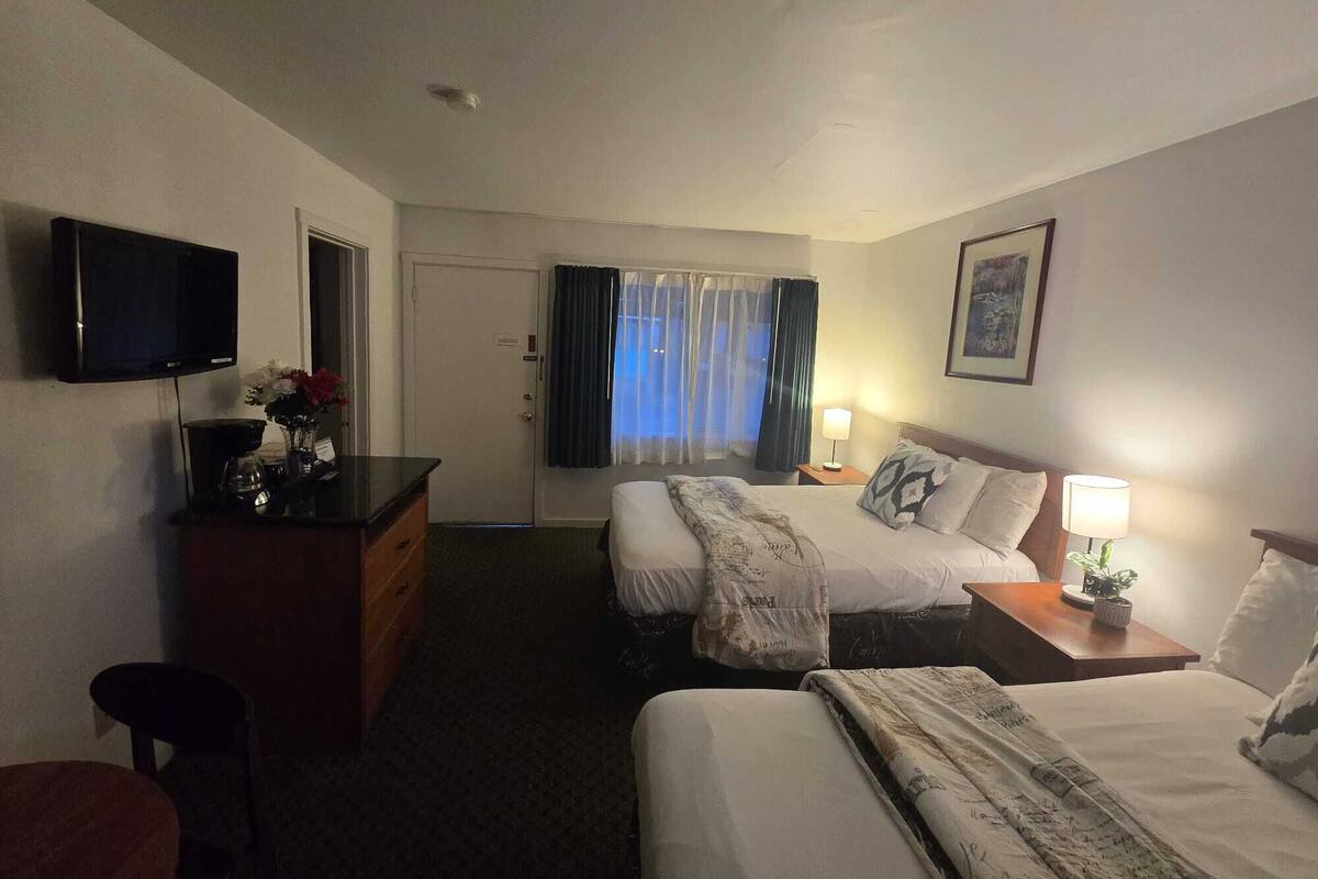 Quarto de hotel com duas camas, televisão, secretária com luminária e janela com cortinas.