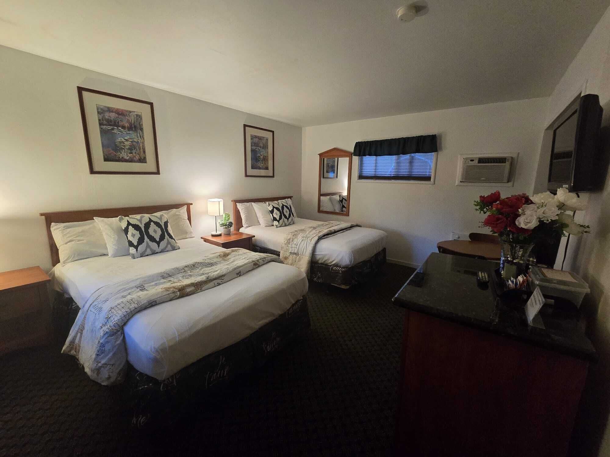 Quarto de hotel com duas camas, uma escrivaninha, um espelho, uma janela com persianas e um ar condicionado fixado na parede.