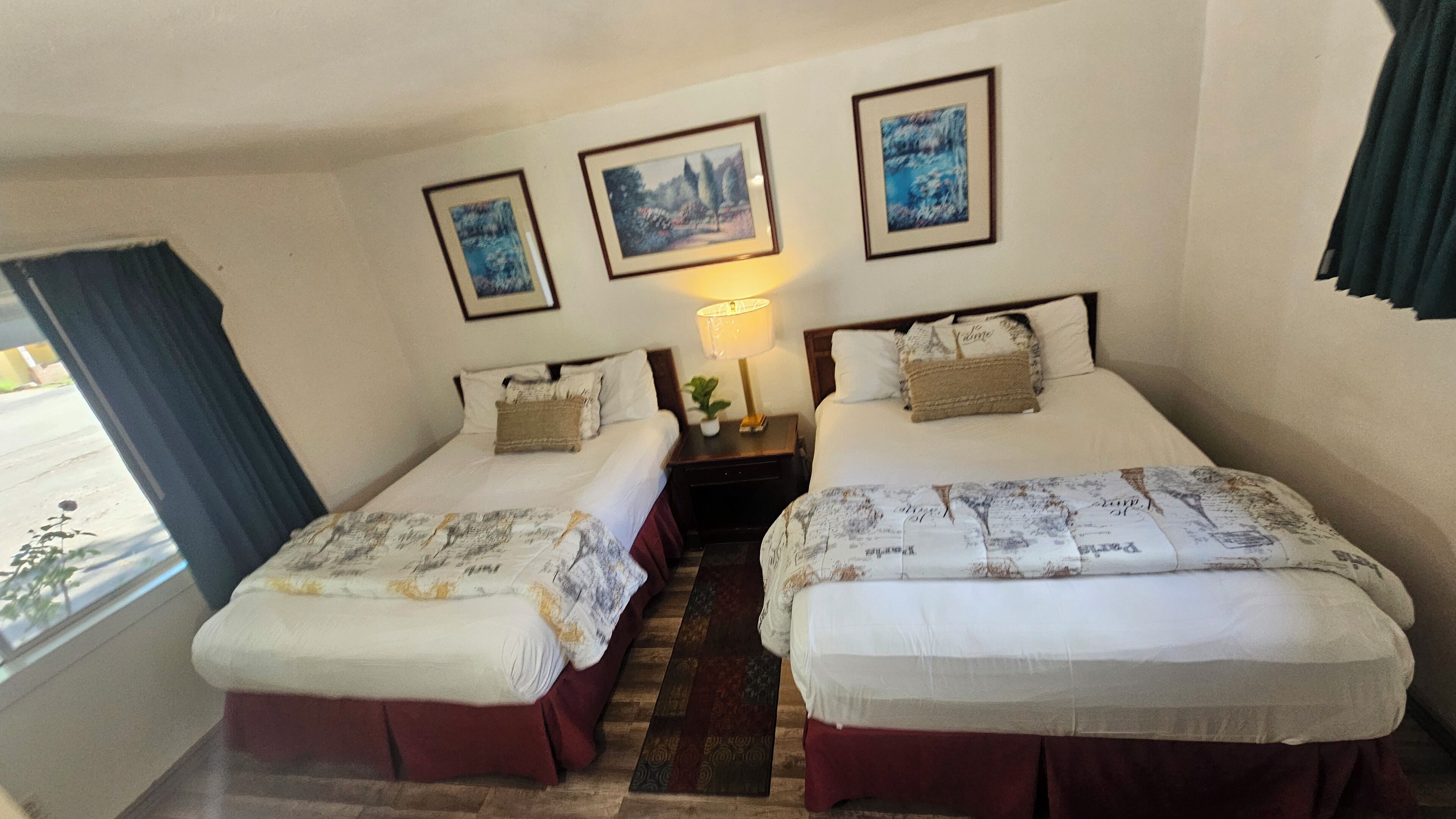 Una habitación de hotel con dos camas, una mesita de noche con lámpara y tres cuadros en la pared.