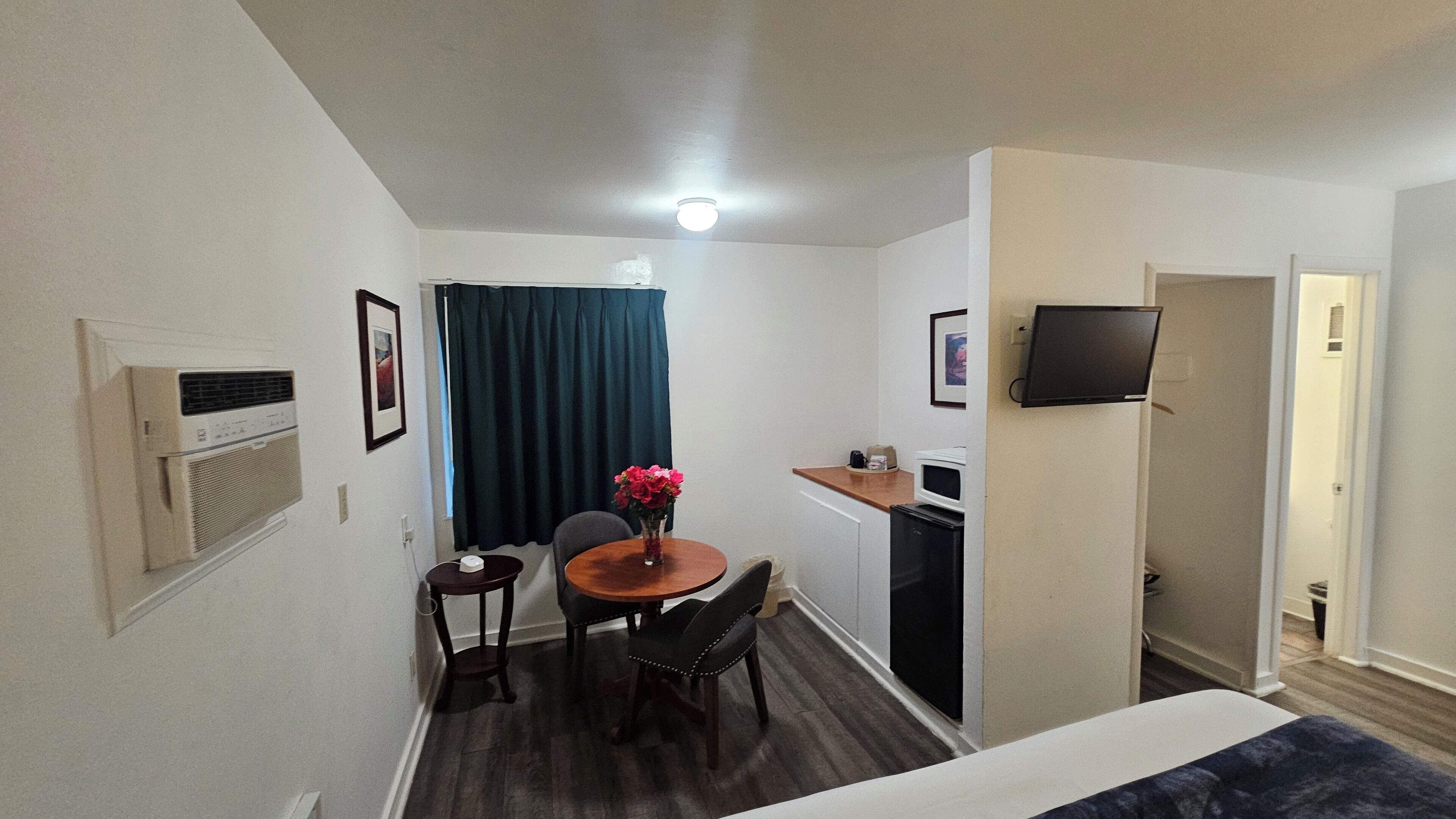 A compact hotel room with a bed, a small kitchenette, a dining area with a table and chairs, a TV mounted on the wall, and a window with curtains.
