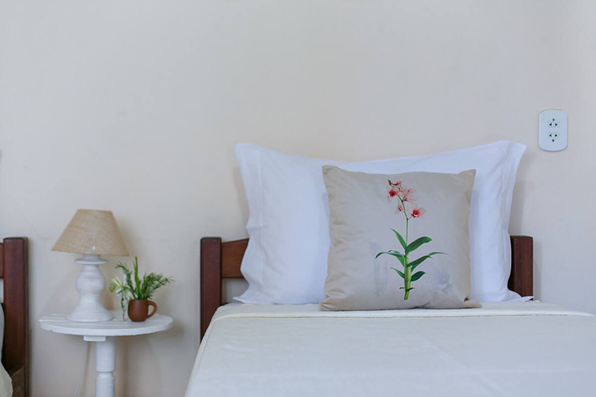 A neatly made bed with white bedding and two pillows, one plain and one with a floral design. A small table with a lamp and a potted plant is beside the bed.
