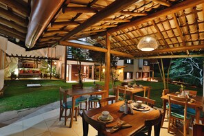 An outdoor dining area with wooden tables and chairs, a pergola with a hanging light fixture, and a view of a house in the background.