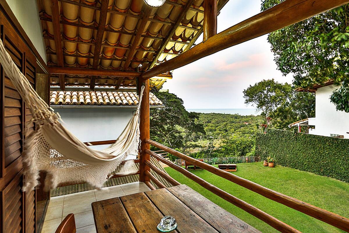 A hammock under a wooden pergola, a wooden table, and a view of a green landscape with a white building in the background.