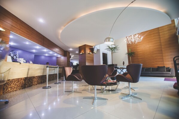 A modern reception area with a wooden reception desk, several seating options, and a decorative wall. - Gontijo Inn Hotel (Itajuba)