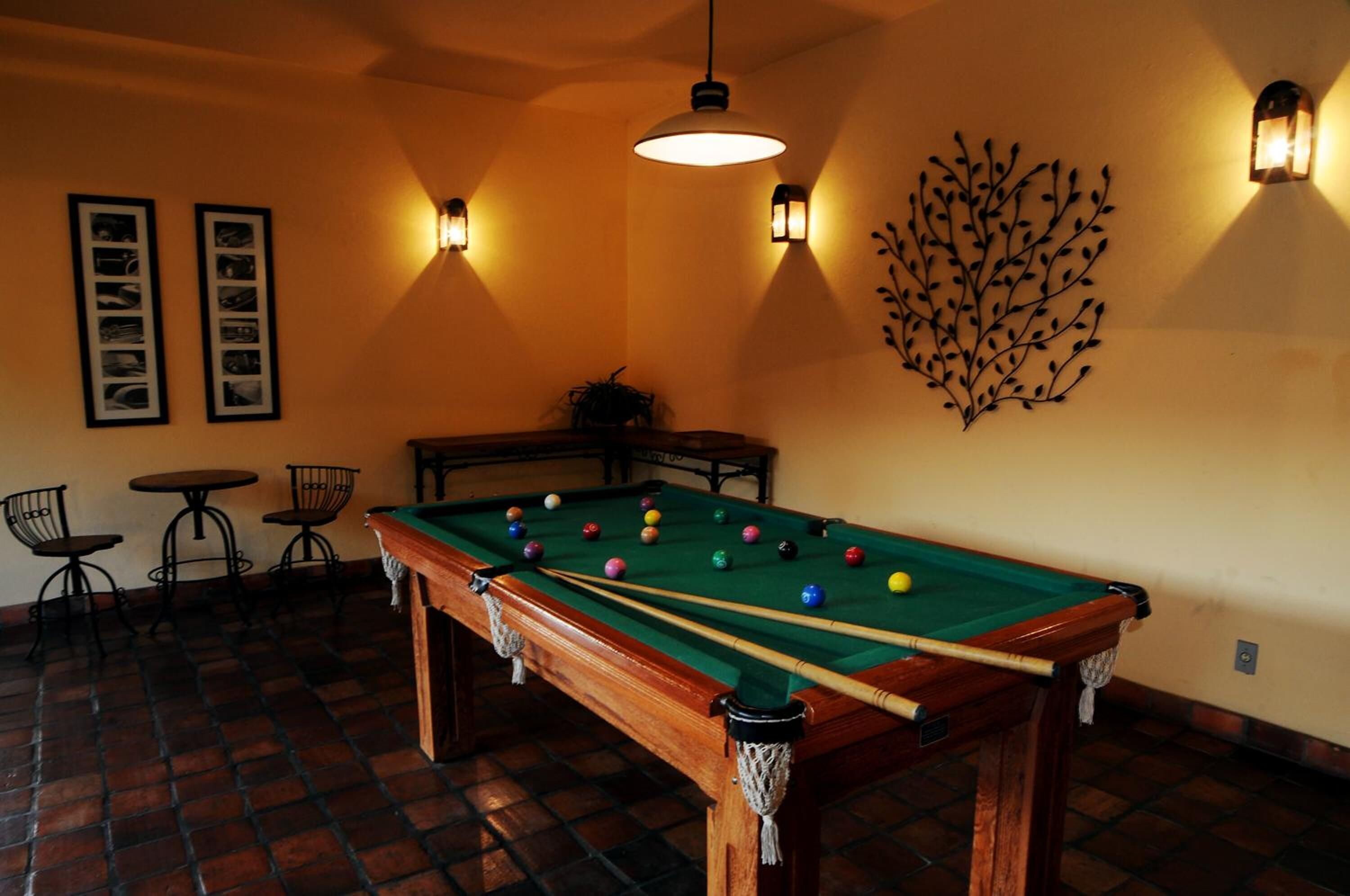 A pool table with balls, two framed pictures, and a decorative wall piece in a room with brick flooring and hanging lights.