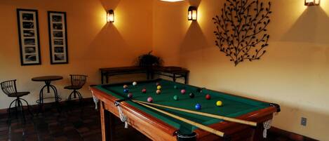 A pool table with balls, two framed pictures, and a decorative wall piece in a room with brick flooring and hanging lights.